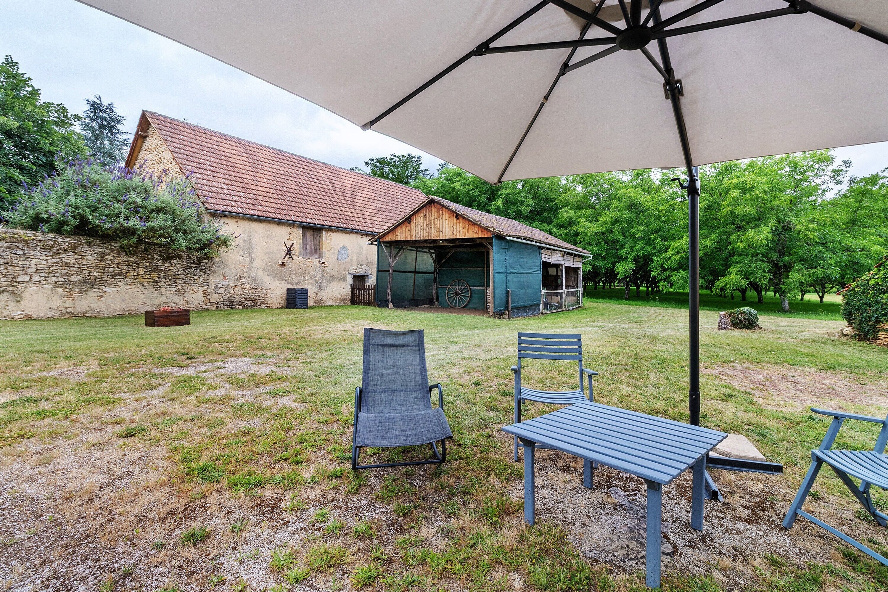 A rustic outdoor seating area with a table and chairs, a large umbrella, and a building with a tiled roof.