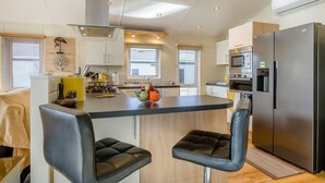 A modern kitchen with a central island, bar stools, a refrigerator, and a microwave. - 3 Bed in Burgh Castle, Great Yarmouth (oc-f34452) (Burgh Castle)
