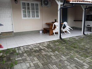 A covered outdoor area with a wooden table and white plastic chairs, a white door, and a window with a grid pattern. - Beach house in Campeche, 1km from the beach. (Florianópolis)