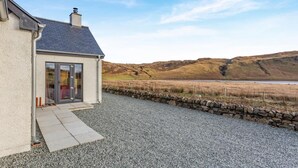 Una casa con camino de grava y muro de piedra, una chimenea, y vistas a colinas ondulantes y un lago.