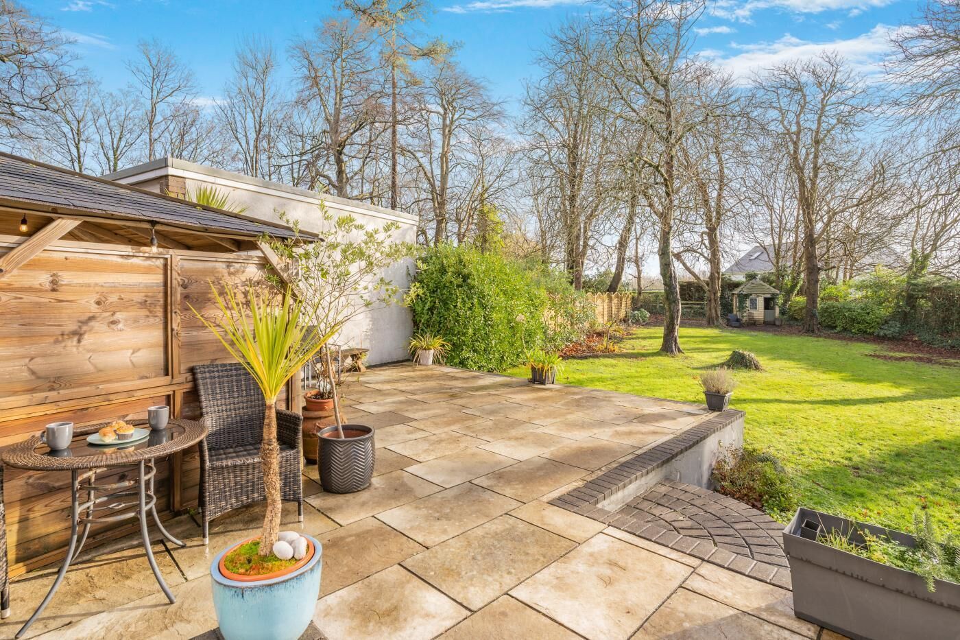 A patio with a wooden table and chairs, surrounded by potted plants and a grassy lawn.