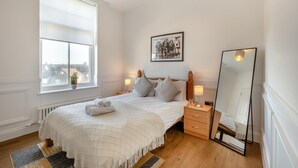 Une chambre avec une grande fenêtre, un lit recouvert d’une couette blanche, une table de chevet avec une lampe, un miroir et un tableau encadré accroché au mur.