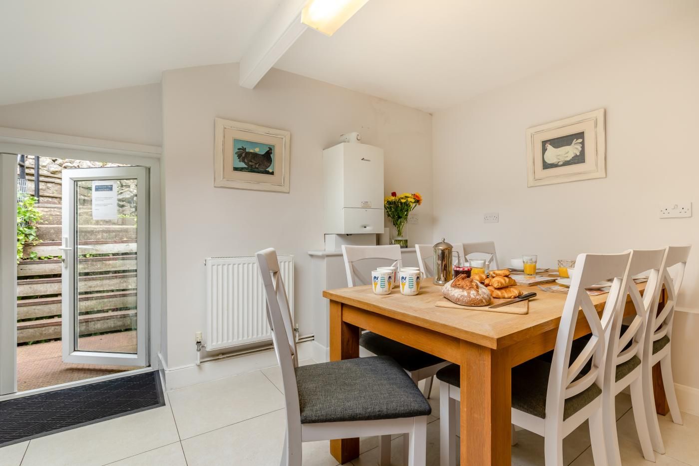 A dining area with a wooden table, white chairs, and a radiator. There are framed pictures on the wall, and a door leading outside.