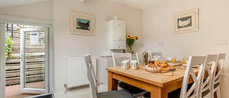 A dining area with a wooden table, white chairs, and a radiator. There are framed pictures on the wall, and a door leading outside.