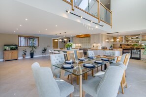A spacious dining area with a glass table, light grey upholstered chairs, and a modern kitchen in the background. - 5 Bed in Spean Bridge (oc-t34246) (Highbridge)