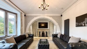 Un salon luxueux avec une cheminée, deux canapés en cuir noir, un pouf capitonné et un lustre.