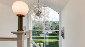 Un escalier avec une rampe en verre, un luminaire suspendu et une vue sur un paysage verdoyant par le biais d’une grande fenêtre.