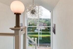 A staircase with a glass railing, a hanging light fixture, and a view of a green landscape through a large window. - 6 Bed in Chapel-en-le-Frith (oc-c34371) (Chapel-en-le-Frith)