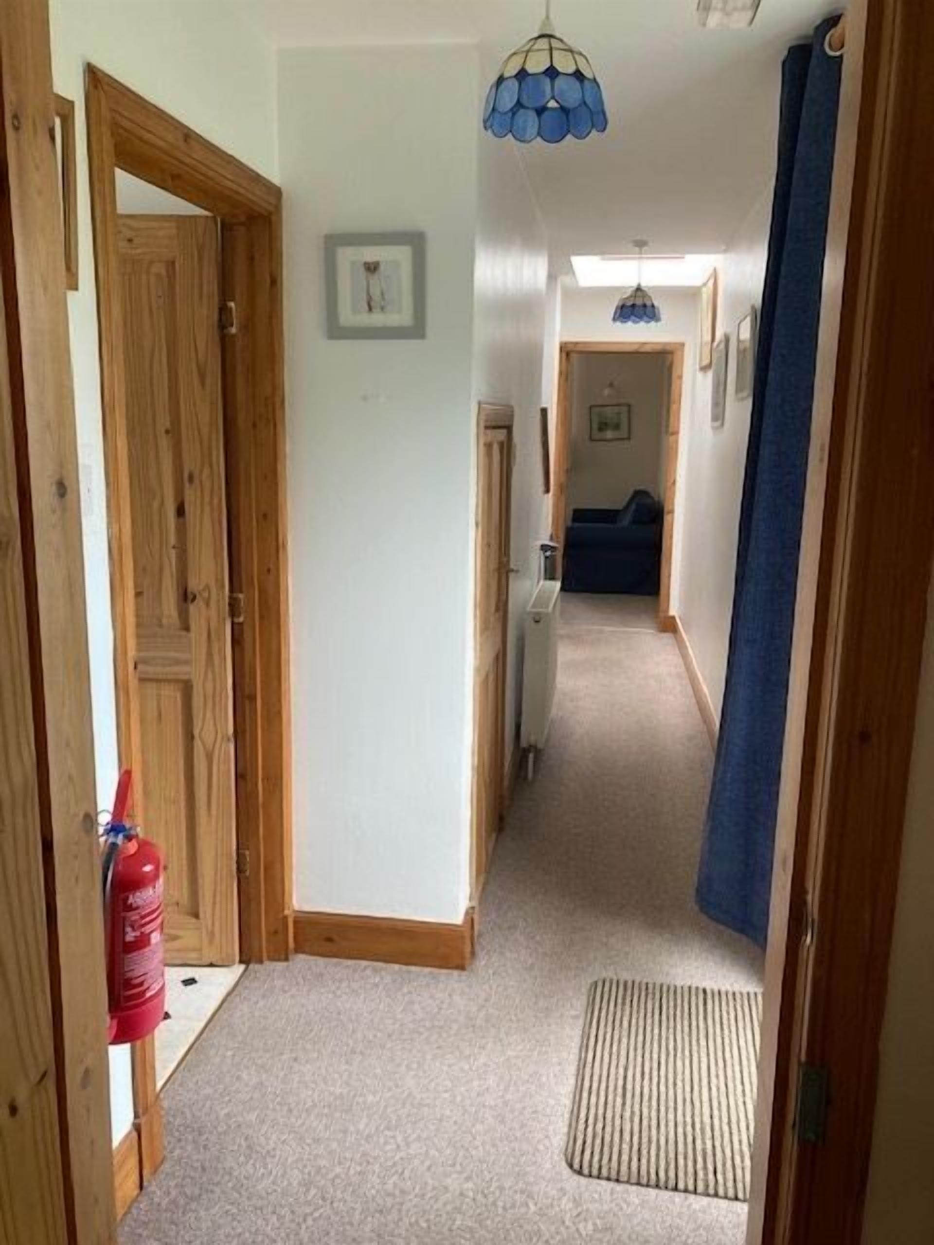 A hallway with wooden doors, a fire extinguisher, a blue hanging light fixture, and a carpeted floor.