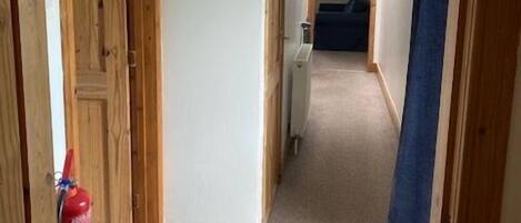 A hallway with wooden doors, a fire extinguisher, a blue hanging light fixture, and a carpeted floor.