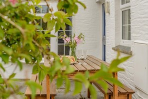 A wooden table with benches, a vase with pink flowers, and a window with a view of a white building. - 3 Bed in St. Merryn (oc-l34453) (Porthcothan Bay)