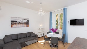 A modern living room with a sectional sofa, a dining table with chairs, a chandelier, a flat-screen TV, and a window with curtains. - Rental in Rome Karan's Suite Apartment (Rome)