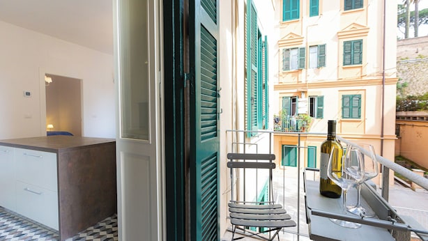 A balcony with a wine bottle and glasses, a chair, and a view of a building with green shutters. - Rental in Rome Karan's Suite Apartment (Rome)