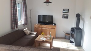 Ett vardagsrum med en soffa, ett soffbord med böcker och blommor, en tv, en öppen spis och rutiga gardiner.