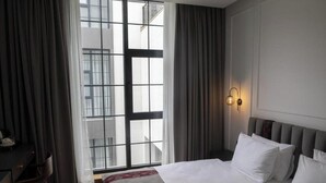 A hotel room with a large window, a bed with white linens, a desk with a chair, and a wall-mounted light fixture. - Molokan Inn Hotel (Baku)