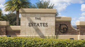 Un cartello con su scritto "THE ESTATES" e un logo, circondato da vegetazione e una palma.