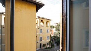 Une vue à travers une fenêtre d’un bâtiment jaune, doté de fenêtres en arc de cercle et donnant sur une cour pavée.