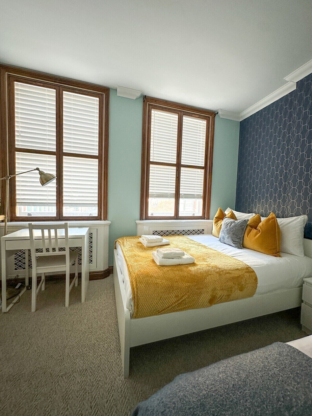 A bedroom with a bed, a desk, and two windows with blinds.