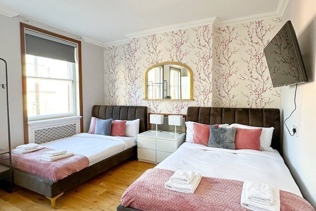 Une chambre avec deux lits, un grand miroir, une table de chevet avec des lampes et une fenêtre avec des stores.