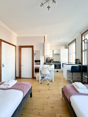 A hotel room with two beds, a desk, a chair, a kitchenette, and a bathroom visible through an open door.