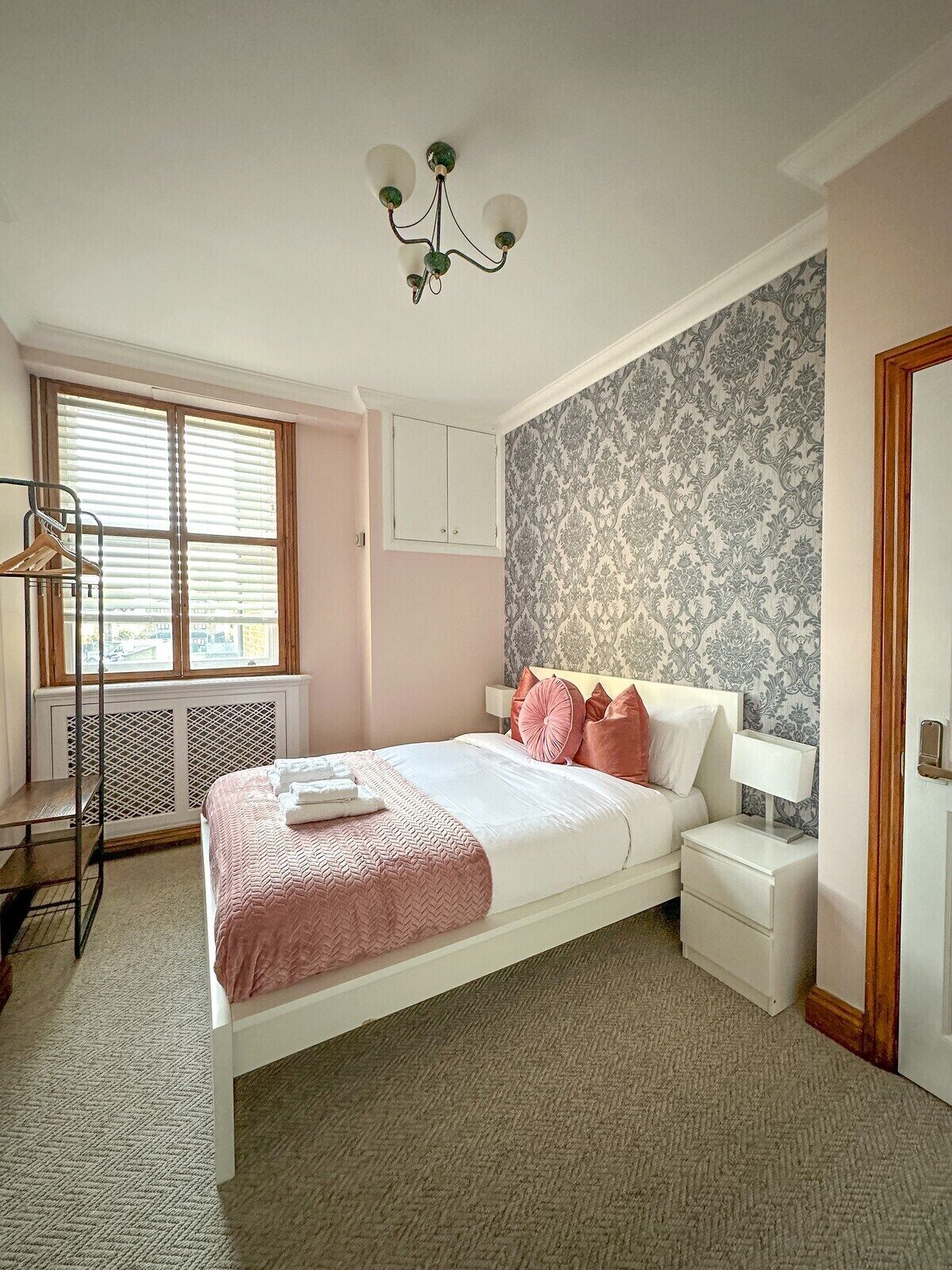 A bedroom with a bed, bedside tables, a radiator, a window with blinds, a wardrobe, and a chandelier.