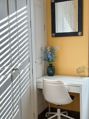 A white desk with a vase of flowers, a mirror, and a chair in a room with a yellow wall and a white door with diagonal blinds. - Fulham Rooms (London)
