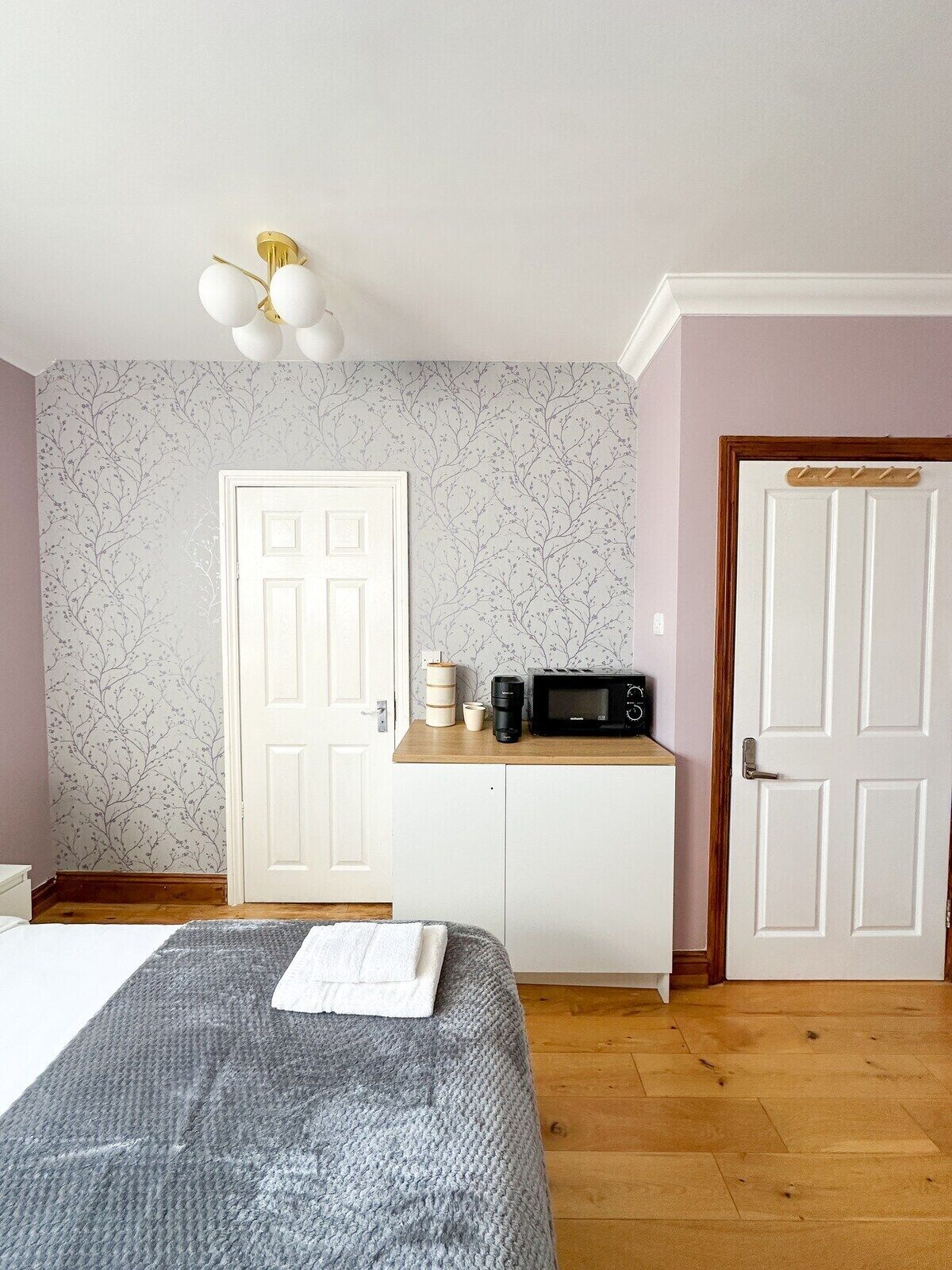A bedroom with a bed, a bedside table with a coffee maker, a microwave, and a door with a coat rack.