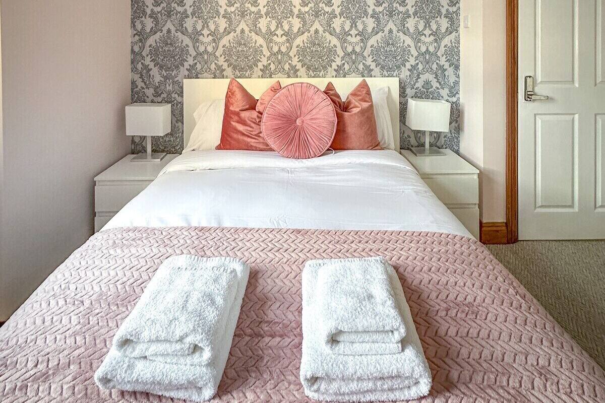 Un lit bien fait, recouvert d’une couette rose et blanche, deux serviettes blanches pliées posées sur le lit, et un papier peint à motifs sur le mur.