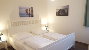 A white bed with two pillows, a nightstand with a lamp, and two wall-mounted pictures. - Apartment 'Fewo Mühlenstr Mit Terrasse' with Private Terrace, Private Garden and Wi-Fi (Prerow)