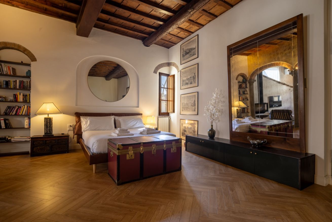 Een slaapkamer met een houten vloer, een bed met hoofdeinde, een nachtkastje, een lamp, een dressoir, een spiegel en lijstjes met foto’s aan de muur.