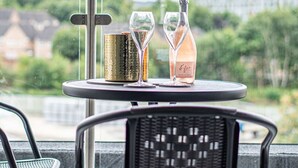 Una mesa con una botella de vino espumoso, dos copas y un recipiente decorativo en un balcón con vistas a la ciudad.