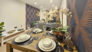 Un comedor con una mesa de madera dispuesta para cuatro personas, rodeada de sillas. Sobre la mesa hay un jarrón grande con flores blancas y una planta en maceta al lado.