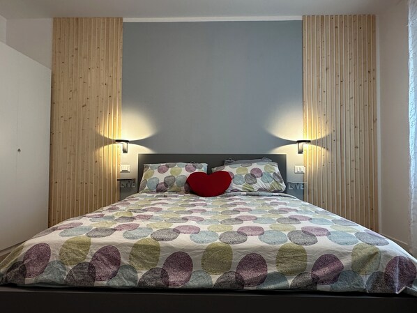 Un letto con una coperta dal motivo circolare colorato, due cuscini e un cuscino a forma di cuore rosso, con due lampade fissate al muro.