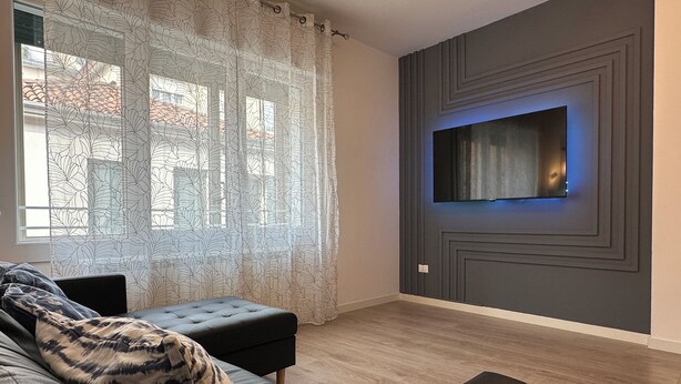 A modern living room with a grey sofa, a patterned throw, a flat-screen TV mounted on a grey wall, and a window with sheer curtains. - [Venice-Mestre]Design Apartment Bros#1 (Venezia)
