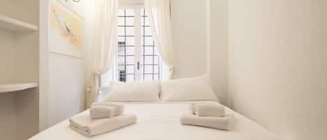 Un lit bien fait, avec du linge de lit et des serviettes blanches, une fenêtre avec des rideaux et un luminaire fixé au mur.