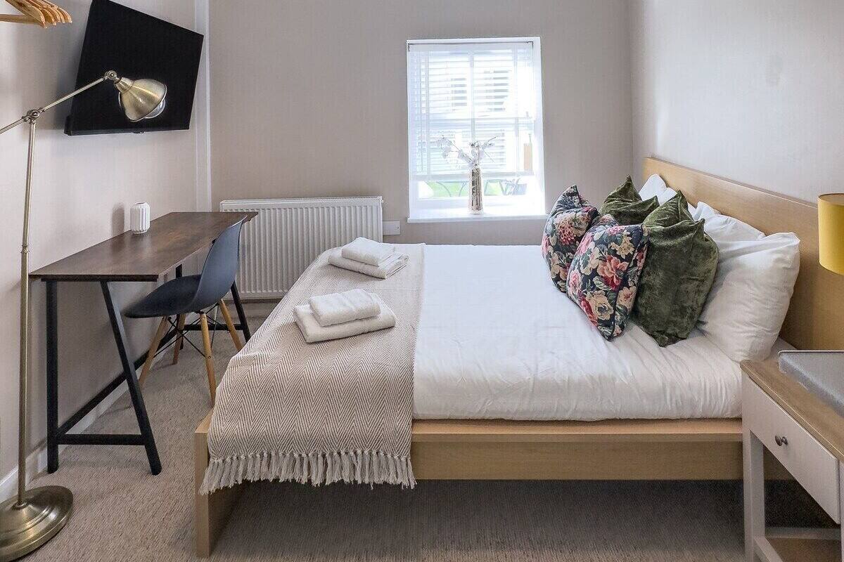 A bedroom with a bed, a desk, a chair, a radiator, a window, and a ceiling light.