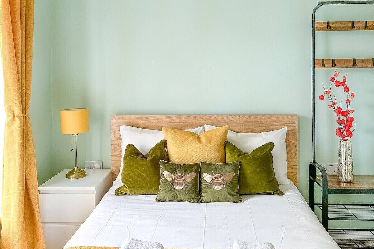 A hotel room with a bed, pillows, a nightstand, a lamp, a mirror, and a vase with flowers.