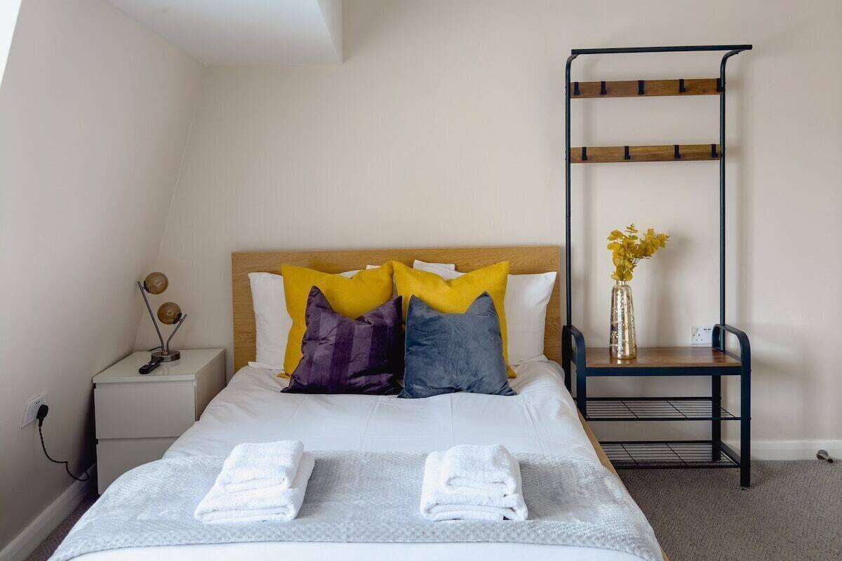 A bedroom with a bed, bedside table, lamp, and a black metal rack with a vase.