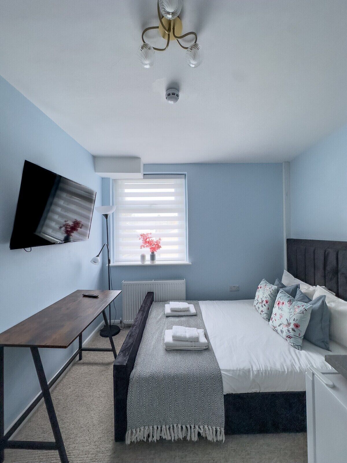A bedroom with a bed, bedside table, lamp, TV, and a window with blinds.