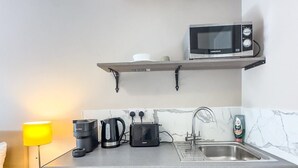 A compact kitchenette with a microwave, toaster, kettle, and coffee maker on the countertop, a sink, and a small refrigerator. - Camden Rooms (London)