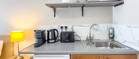 A compact kitchenette with a microwave, toaster, kettle, and coffee maker on the countertop, a sink, and a small refrigerator.