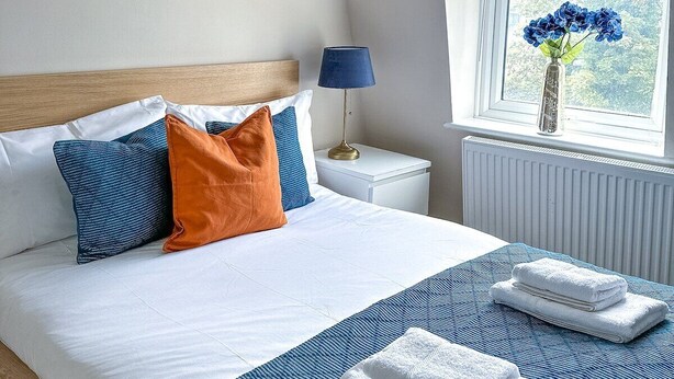 A bedroom with a bed, pillows, a bedside lamp, a window with a view, and a radiator. - Camden Rooms (London)
