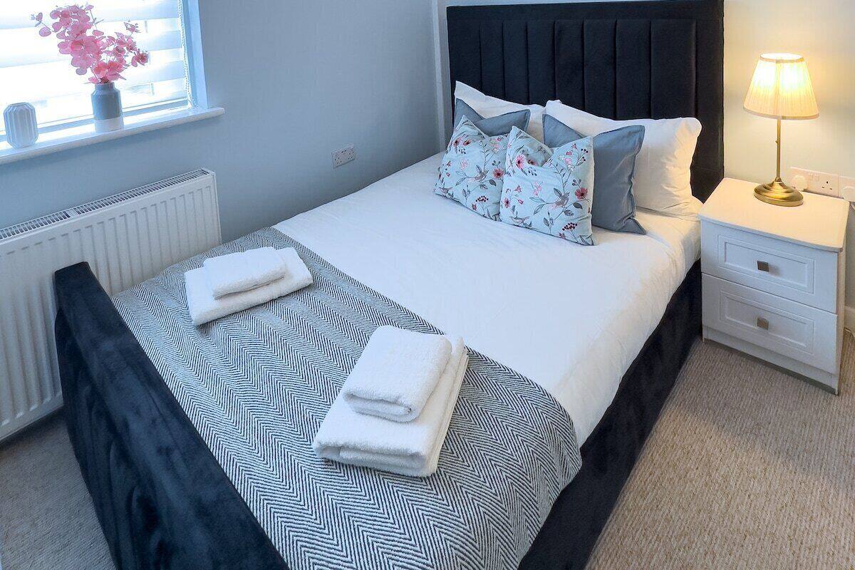 A bedroom with a bed, bedside table, lamp, window with blinds, radiator, and a potted plant.
