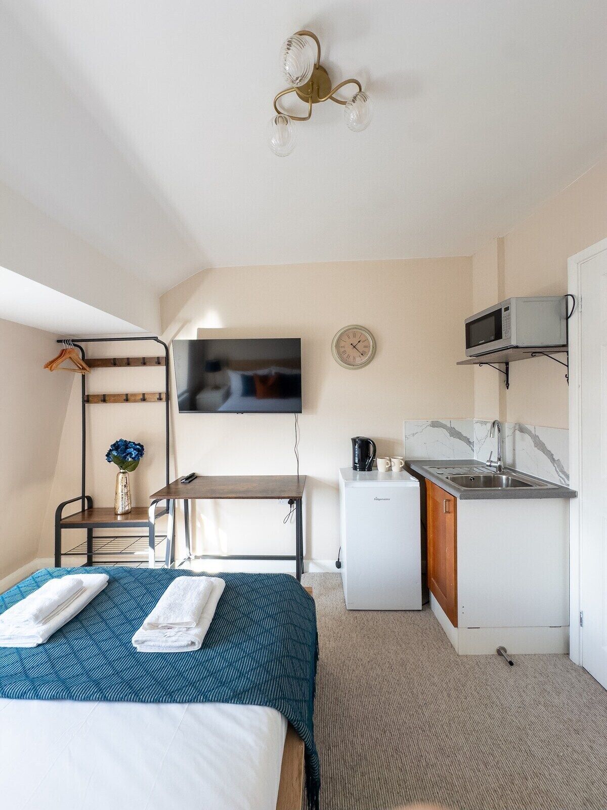 A compact hotel room with a bed, a small kitchenette, a TV mounted on the wall, and a ceiling light fixture.