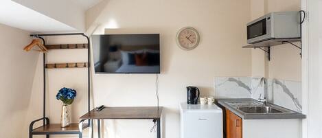 A compact hotel room with a bed, a small kitchenette, a TV mounted on the wall, and a ceiling light fixture.