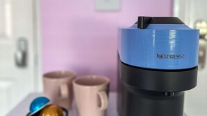 A Nespresso coffee machine with a blue and black body, a coffee pod, and two coffee cups on a table. - Camden Rooms (London)