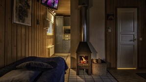 Ein gemütliches Zimmer mit einem Bett, einem an der Wand montierten Fernseher, einem Kaminofen und einer Tür zum Küchenbereich.