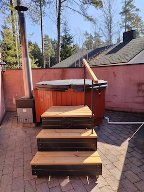 Kylpytynnyri, jonka ympärillä on puinen terassi ja metallikaide. Tynnyrin ympärillä on tiilipäällysteinen patio ja punainen seinä.