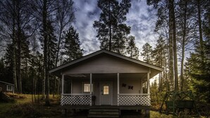 Une cabane dotée d’une véranda, entourée de grands arbres et surplombée par un ciel nuageux.