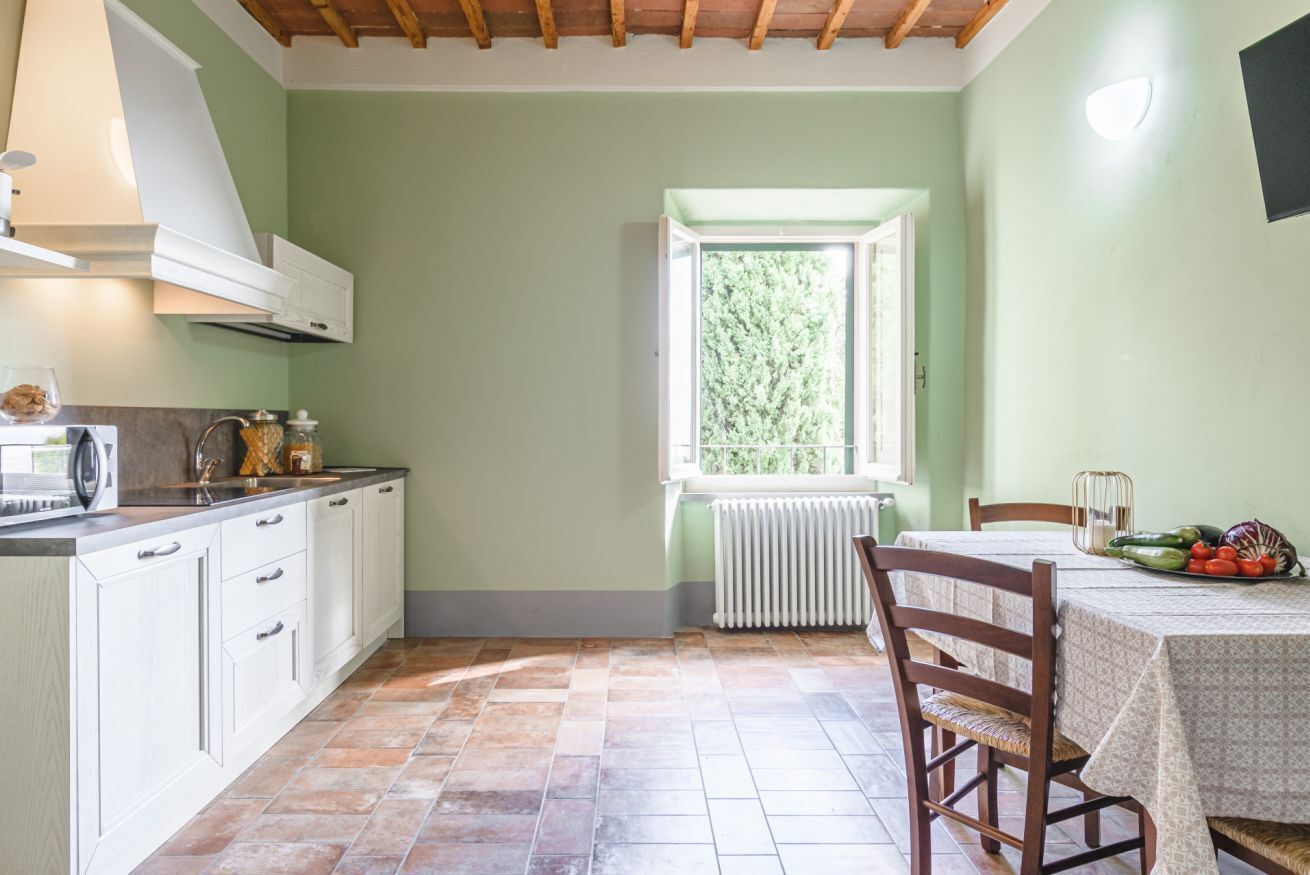 Cocina con armarios blancos, una ventana con vistas a zonas verdes, y una mesa con mantel y verduras.